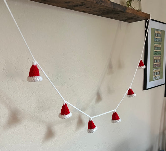 shows a tiny crochet santa hat garland garland. the santa hats are red with a white brim and pom pom, and connected with white yarn. the garland is shown hanging from a shelf against an off-white wall background.